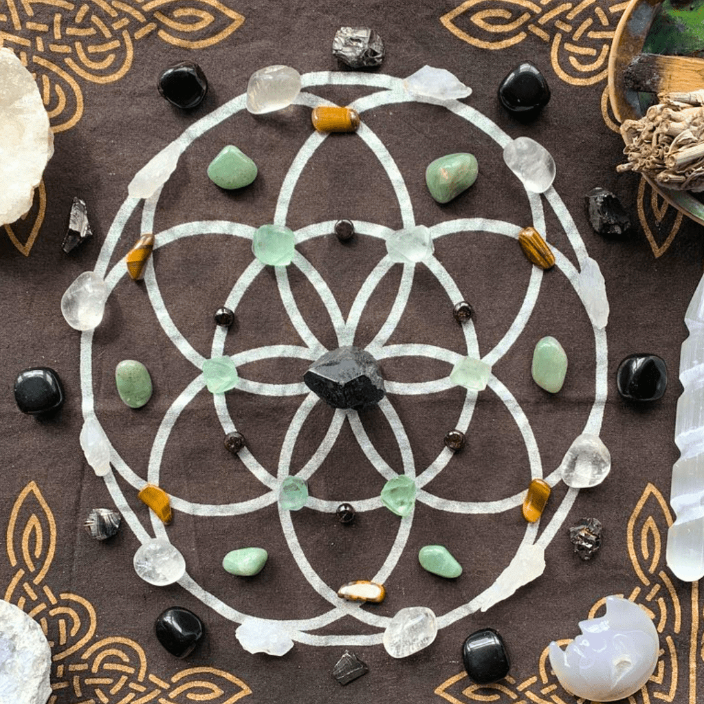 An image of a crystal grid laid out on a black cloth with white geometric patterns representing the Seed of Life. At the centre of the grid is a large, rough black stone, likely Black Tourmaline. Radiating outwards in concentric circles are various crystals: fluorite, garnet and clear, milky stones that are quartz  positioned in the innermost circle, followed by a mix of green and brownish stones, perhaps Green Aventurine and Tiger's Eye, in the middle layer. The outermost circle contains dark, polished stones, potentially Black Obsidian. Additional small, dark stones, and a translucent pointed crystal are placed at the grid's periphery. To the side, there's a smudge stick in a shell and a chunky, clear crystal cluster.