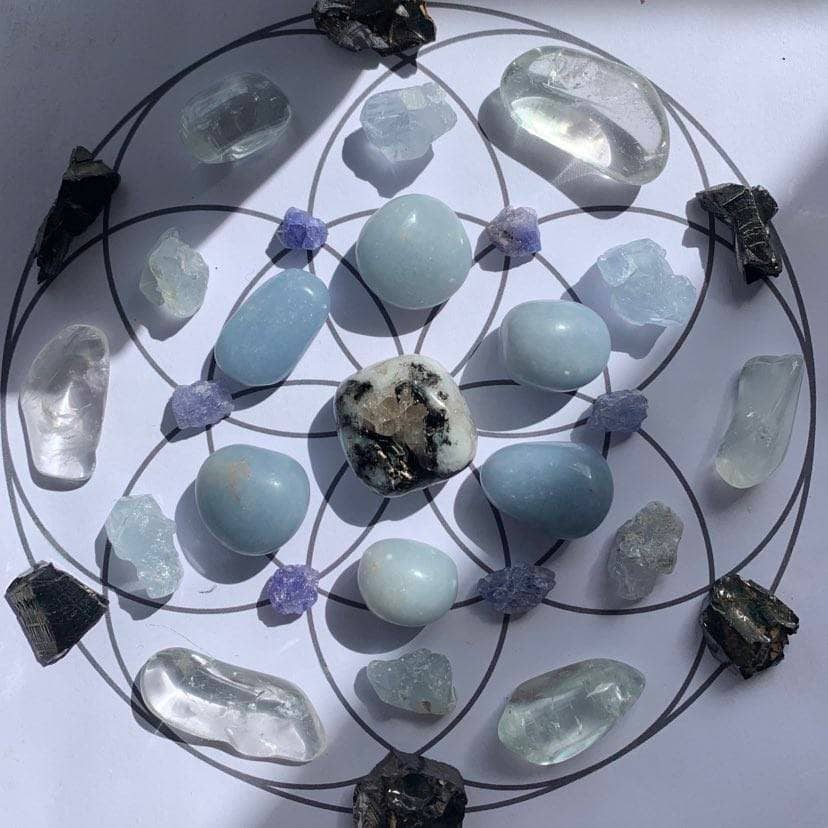This image shows a crystal grid for the first quarter moon in Cancer, arranged on a circular template with interconnected circles, resembling a flower or geometric pattern. At the centre, there is a large, prominent Rainbow Moonstone, which is known for its connection to intuition and the Moon. Radiating outward are smooth, polished Angelite stones, which have a soothing blue colour, enhancing the nurturing energy of the grid. Interspersed between them are smaller, rough Tanzanite crystals, their bluish-purple tones adding a touch of transformational energy. Bordering the grid are shards of dark, reflective Shungite, which contribute grounding and protective properties. Completing the perimeter are clear, translucent points of Praseolite, which can amplify the energies of the other stones. The overall arrangement and selection of crystals seem intentional, aiming to create a harmonious and powerful energetic field.