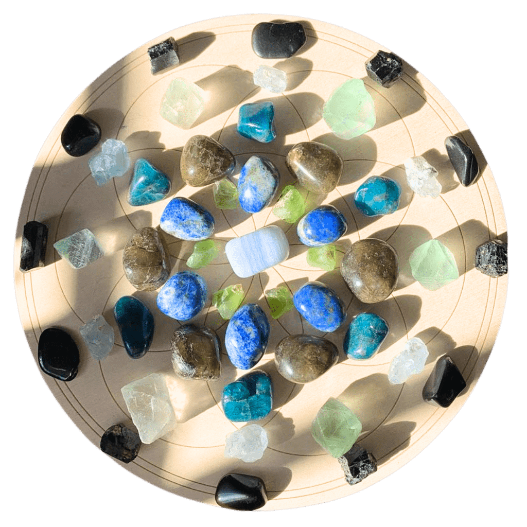 A circular wooden crystal grid featuring an arrangement of various tumbled and raw crystals, including blue lace agate, lapis lazuli, labradorite, peridot, fluorite, and black tourmaline, placed in a symmetrical pattern.